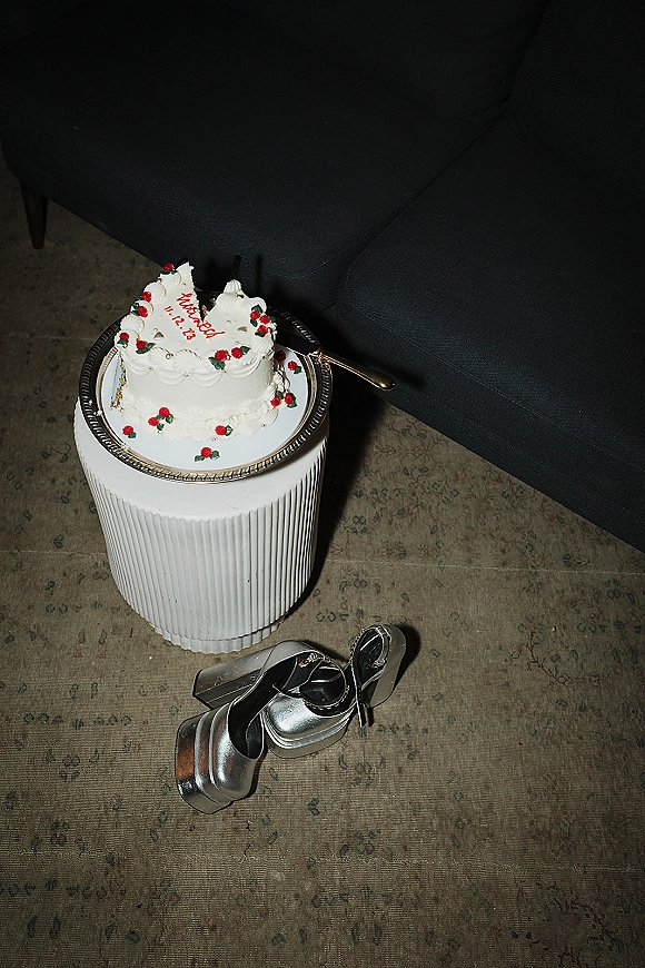 Wedding cake shaped like a heart with white frosting, cherry decorations, and piped message on a pedestal beside silver heels