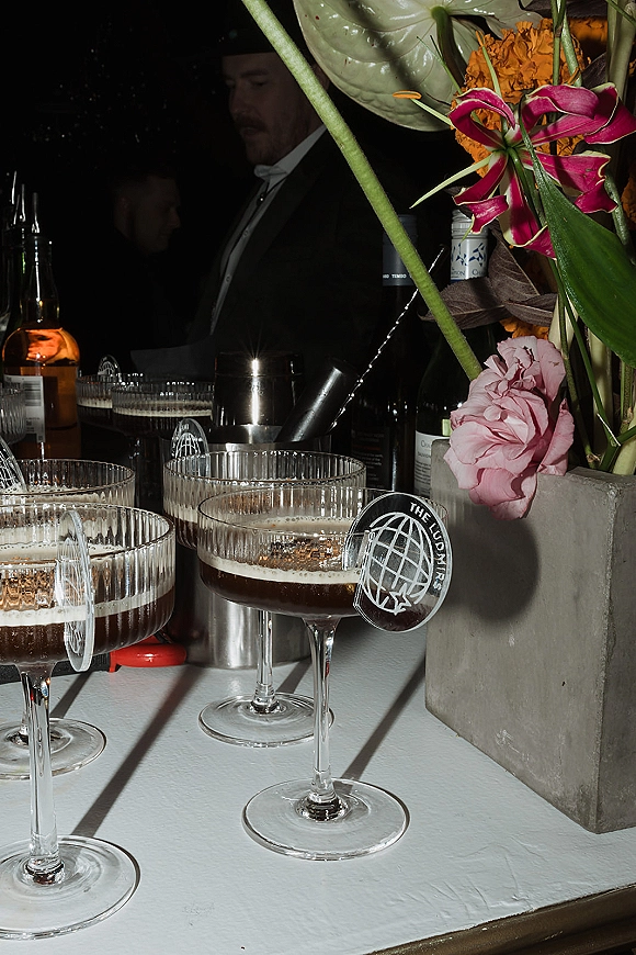 Wedding cocktail bar with wedding signature cocktails in coupe glasses, wine bottles, and a concrete vase floral centerpiece on a white table