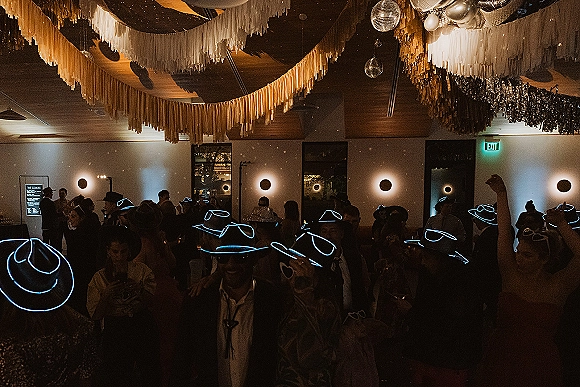 Wedding dance floor packed with guests in glow stick party hats under fringe ceiling decor, disco balls, and string lights in a dark reception room