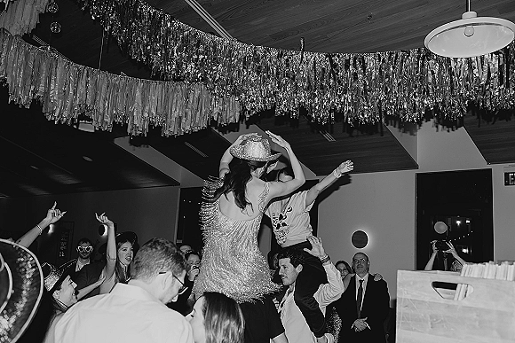 Wedding dance party with a hora chair lift as bride and groom are hoisted above cheering guests under tinsel garland in a reception hall