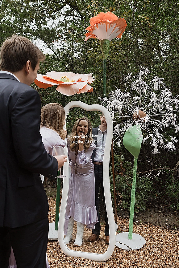 Wedding photo booth guests in suits and a long-sleeve dress pose with oversized flower props by a standing mirror on a garden gravel path