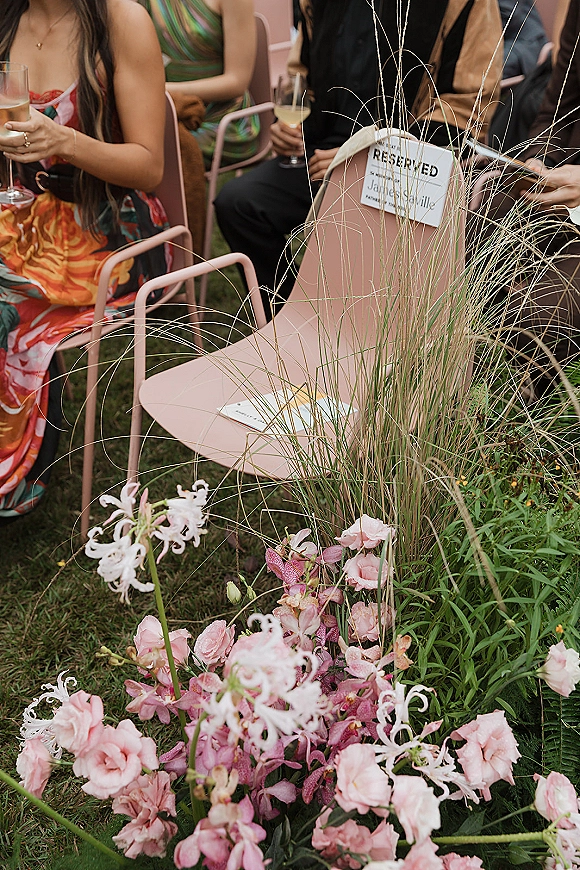 Ceremony seating with a reserved seat sign on pink metal chairs, champagne flutes nearby, and floral ground arrangement on a grass lawn