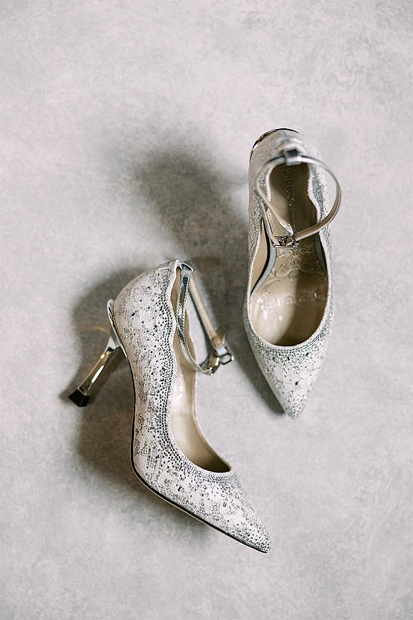 Bridal shoes with sparkly wedding heels detail, rhinestone accents and lace overlay on ankle straps, resting on light gray fabric flat lay