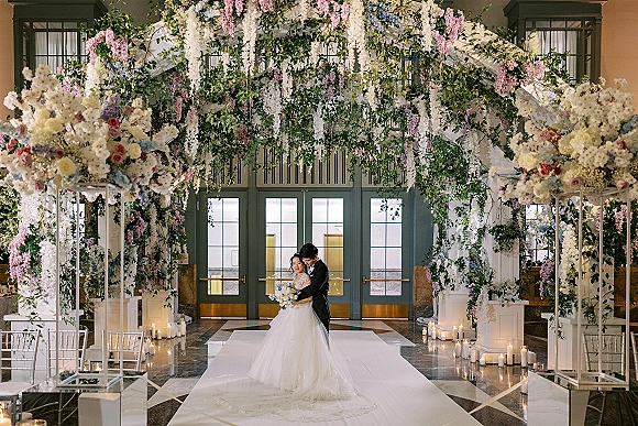 Couple portrait of bride and groom embrace beneath an indoor wedding arch with hanging florals, candles, and marble floor backdrop