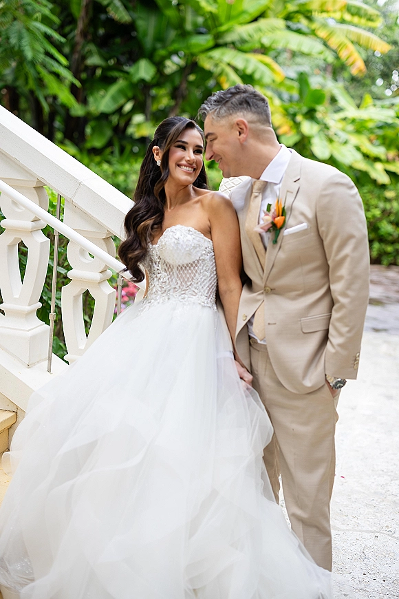 Couple portrait of bride and groom leaning together, her strapless ballgown and his suit on white stairs amid tropical greenery outdoors