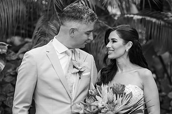 Couple portrait of bride and groom smiling close together, bride holding a bouquet in a strapless lace dress against palm leaves.