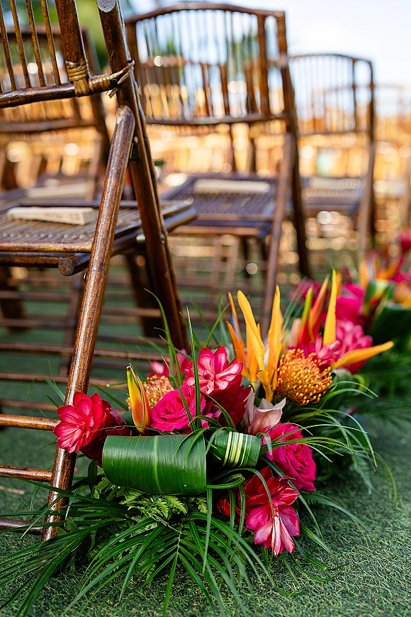 Ceremony aisle decor with tropical aisle flowers and palm leaves beside wood folding chairs on an outdoor lawn under a blurred sky