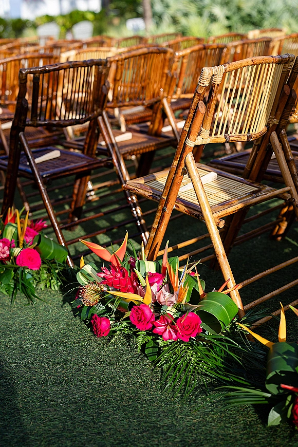 Ceremony seating with bamboo wedding chairs in sunlit garden rows, lined with tropical floral arrangements of roses and bird of paradise
