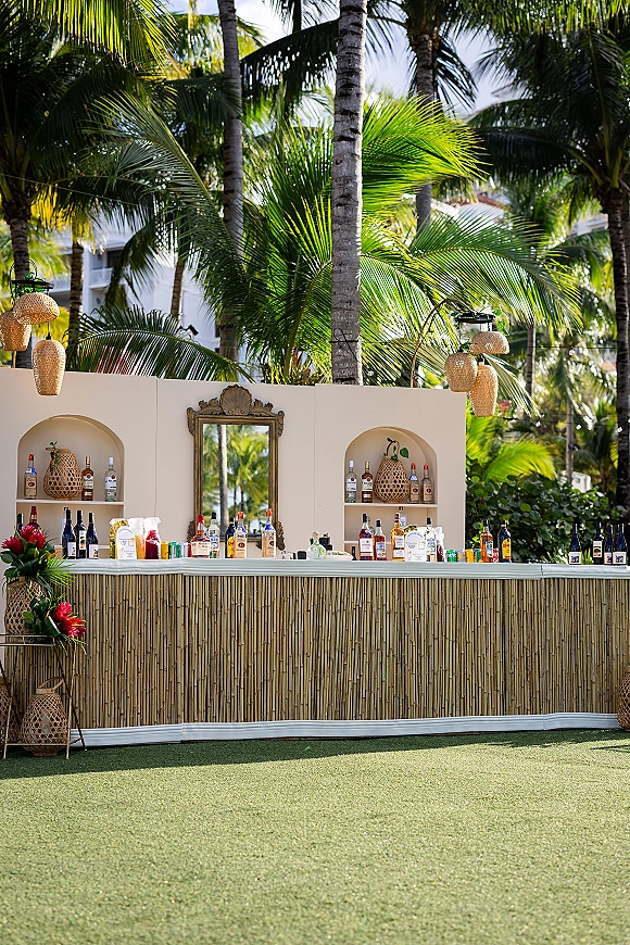 Outdoor bar setup with wedding bar decor, bamboo front, liquor bottles and mirror backdrop under rattan pendant lights on a palm-lined lawn