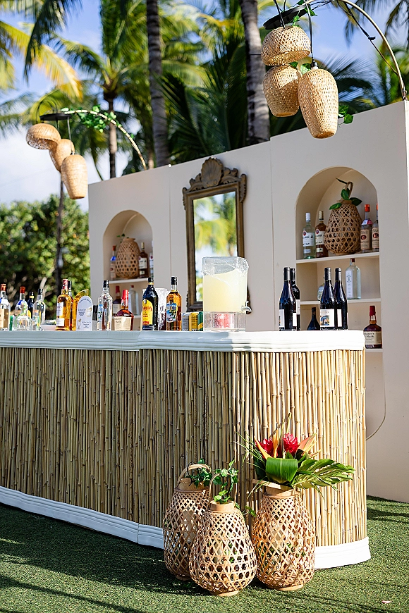 Outdoor wedding bar with bamboo front, tiki wedding bar shelving of liquor and wine bottles under rattan lights on a palm-lined lawn