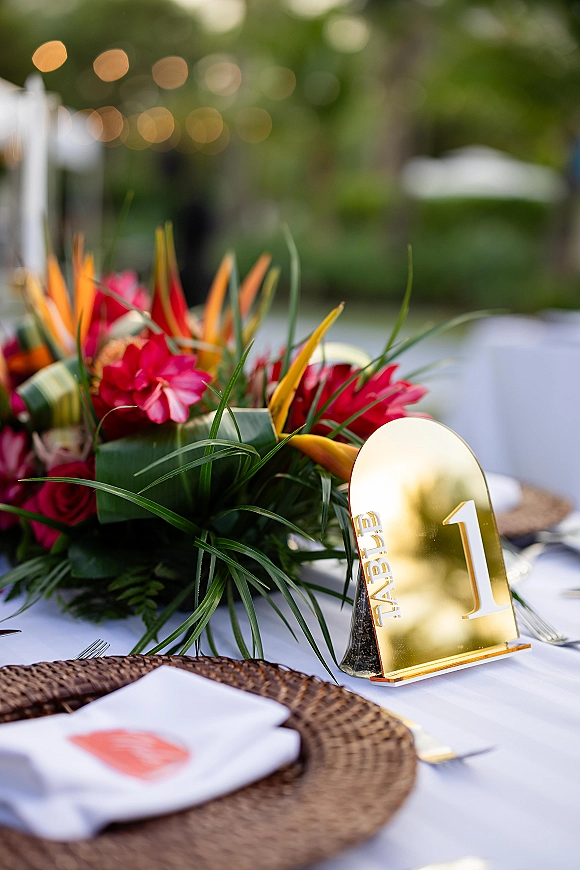 Reception tablescape with tropical wedding centerpiece of bird of paradise and red flowers, gold table number, string lights bokeh outdoors