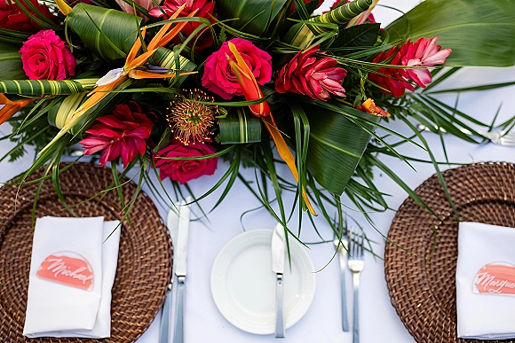 Reception tablescape with tropical wedding tablescape flair, featuring a tropical floral centerpiece of hot pink roses, bird of paradise, and palm leaves