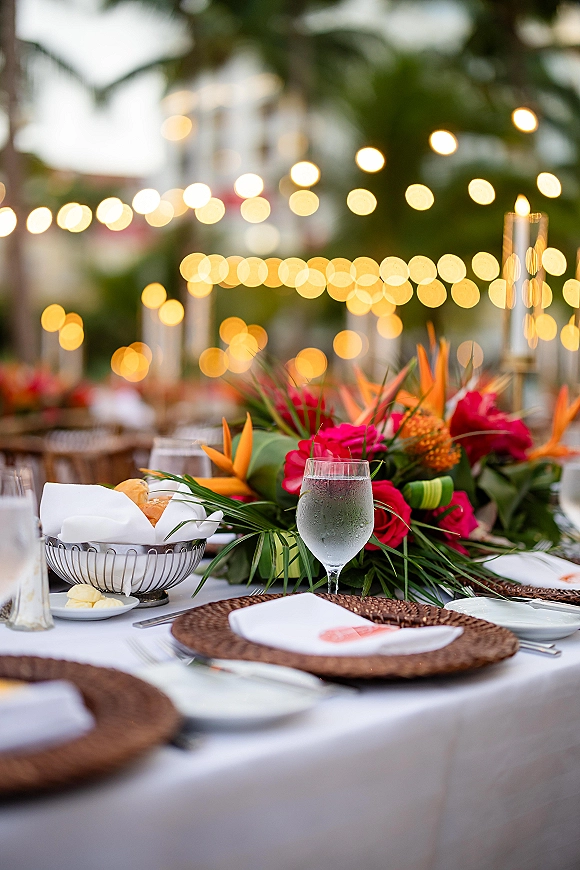 Reception tablescape with an outdoor reception table featuring bird of paradise and pink roses, candle hurricanes, and string lights under palms at dusk