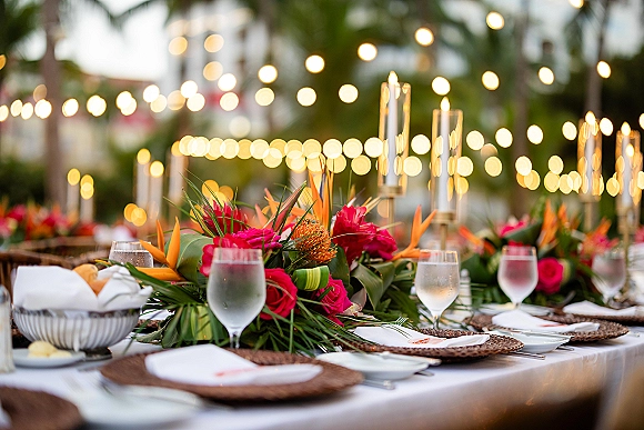 Reception tablescape with tropical wedding tablescape florals, birds of paradise, pink roses, taper candles and string lights under palms at dusk