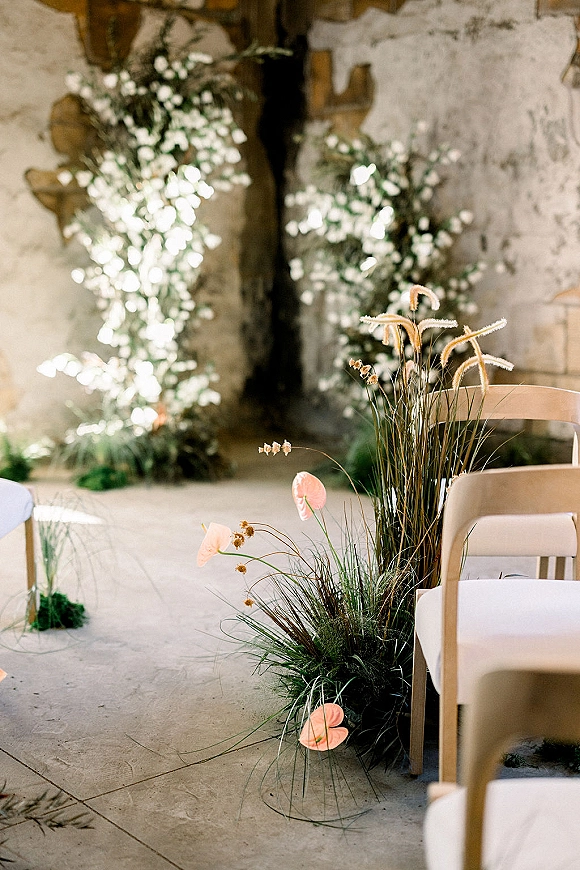 Ceremony aisle decor with wedding aisle flowers in low white blooms, greenery and pink anthurium along chairs beside a stone wall