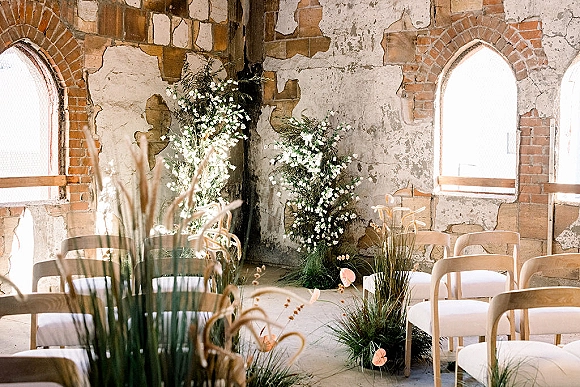 Ceremony setup with white floral arrangements and greenery, pampas accents framing a meadow grass aisle in an industrial room with arched windows