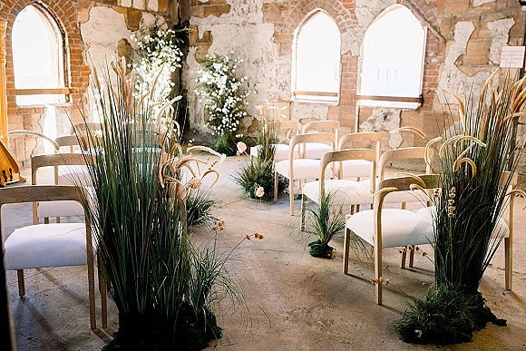 Ceremony setup with white aisle flowers and pampas grass lining wooden chairs in a natural light hall with arched windows and brick walls