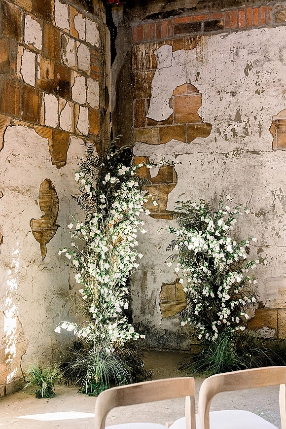 Ceremony backdrop with wedding ceremony backdrop flowers in white and greenery, set against a distressed plaster and brick wall with chairs