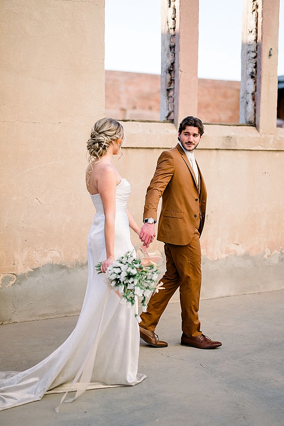 Couple portrait of bride and groom holding hands, bride in strapless dress with greenery bouquet, walking by a stucco wall outdoors