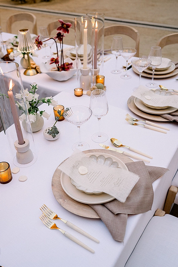 Reception tablescape with wedding place setting, scalloped plates, gold flatware, coupe glasses, taper candles and bud vases on stone floor