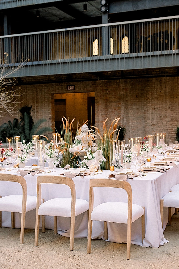 Reception tablescape with a round wedding reception table, white linens, candlelit floral centerpieces and wedding cake against a brick wall backdrop