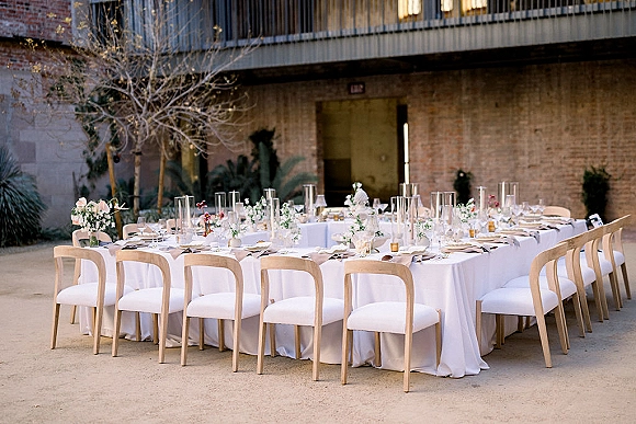 Reception tablescape with long wedding table setup in white linens, greenery and floral centerpieces, taper candles, and brick courtyard backdrop