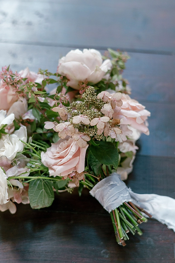 Bridal bouquet of blush roses and white blooms with greenery and ribbon wrap, resting on a dark wooden surface in soft light