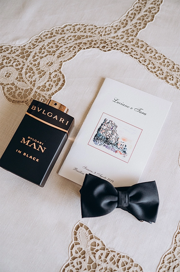 Wedding invitation suite flat lay with illustrated card, black bow tie, and cologne bottle on white linen with lace trim