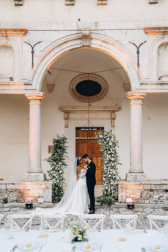 Wedding couple portrait of bride and groom embrace under a stone archway, her cathedral veil and tiara framed by white rose pillars and chandelier