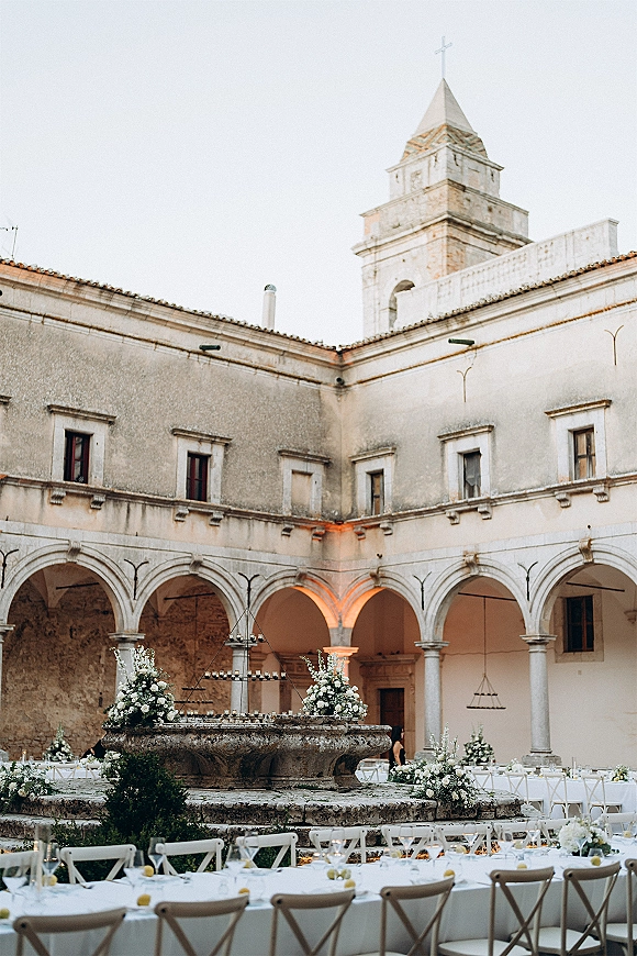Courtyard wedding reception with long banquet tables in white linens, floral centerpieces and candles around a stone fountain in a cloister courtyard