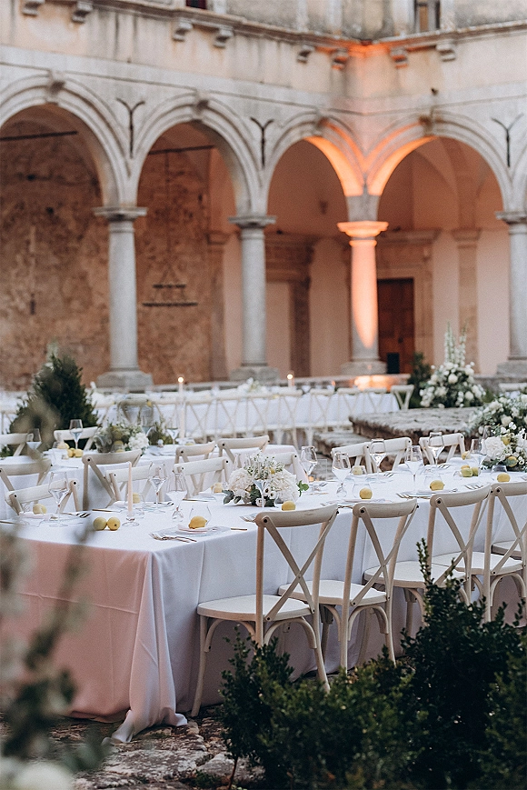 Reception tablescape with long banquet table wedding styling, white tablecloths, floral centerpieces, candles, and lemons in stone courtyard