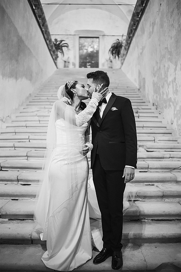 Wedding kiss portrait in black and white of bride and groom kissing on a stone staircase beneath an arched doorway, veil trailing