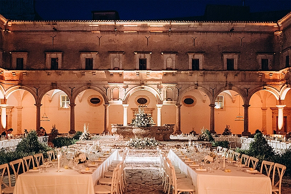 Courtyard reception setup with long banquet tables in white linens, taper candles, and florals around a stone fountain under arched colonnades at night