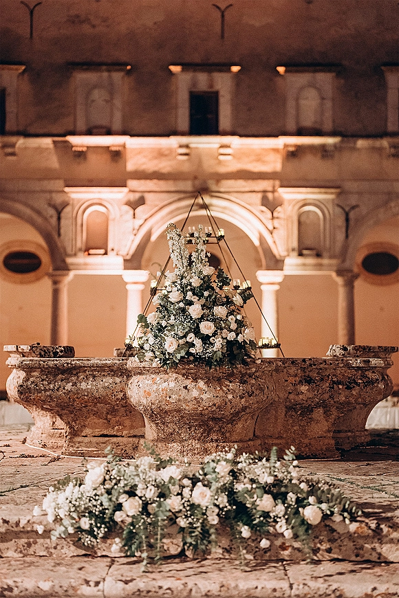 Wedding ceremony decor with ceremony altar flowers, white blooms and eucalyptus, hanging installation, candles, and stone fountains in a historic courtyard with arches