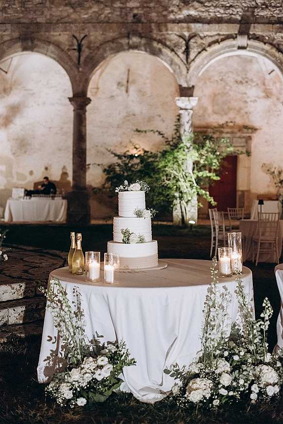 Wedding cake table with a three tier wedding cake, white roses and candles in glass holders, set in a stone-arched courtyard