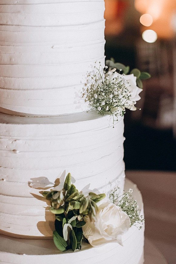 Wedding cake with white buttercream frosting and horizontal ridges, topped with baby's breath, white roses, and greenery against warm bokeh lights