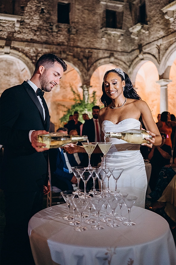 Champagne tower moment as bride in strapless dress and groom in tux pour bottles into coupe glasses under stone arches with warm uplighting