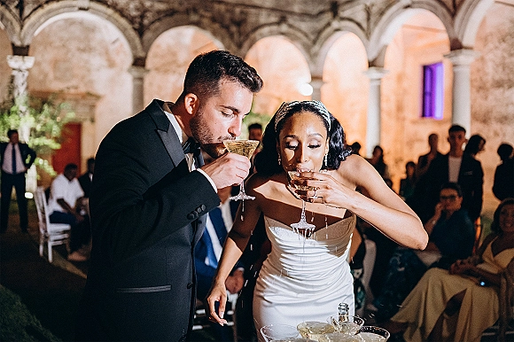 Champagne toast moment as bride and groom toasting with coupe glasses, smiling under stone arches in an uplit indoor courtyard with guests