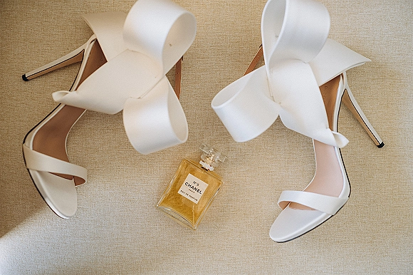 Bridal shoes in a flat lay with white wedding heels featuring oversized bows beside a perfume bottle on neutral fabric background