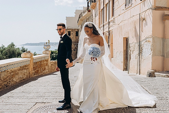 First look moment as bride taps groom’s shoulder on a sunny stone street by the sea, her long veil trailing behind a strapless gown