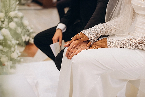 Ceremony moment with bride and groom hands holding a wedding ring close up, lace sleeve and veil accents along an indoor aisle with greenery