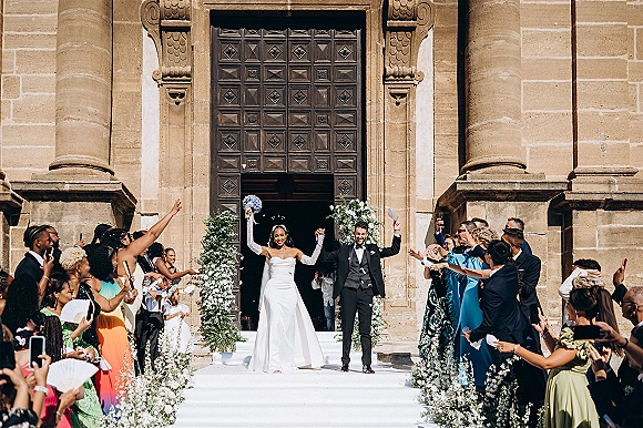 Wedding recessional as newlyweds walking down aisle, bride lifting bouquet beside cheering groom, exiting stone church with guests filming