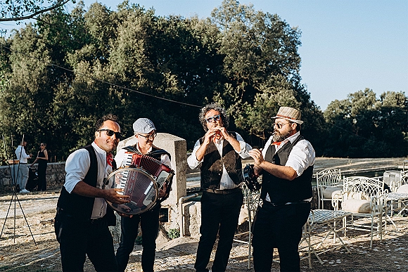 Wedding band playing live wedding music with accordion and tambourine in a sunny stone courtyard with trees and patio chairs
