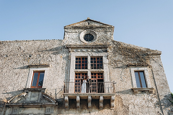 Couple portrait of bride and groom balcony pose in sunglasses, full length by railing against an ivy-covered stone facade and blue sky