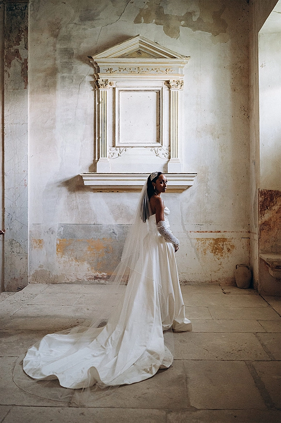 Bridal portrait of a bride with long veil in a strapless gown and lace opera gloves, standing by a textured plaster wall in window light