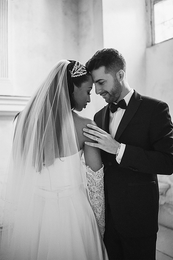 Couple portrait in a black and white wedding portrait, bride and groom forehead touch in close embrace by window light, veil and tuxedo visible