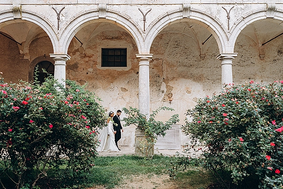 Couple portrait of bride and groom walking side profile, veil trailing, between rose bushes by stone arches in a courtyard setting