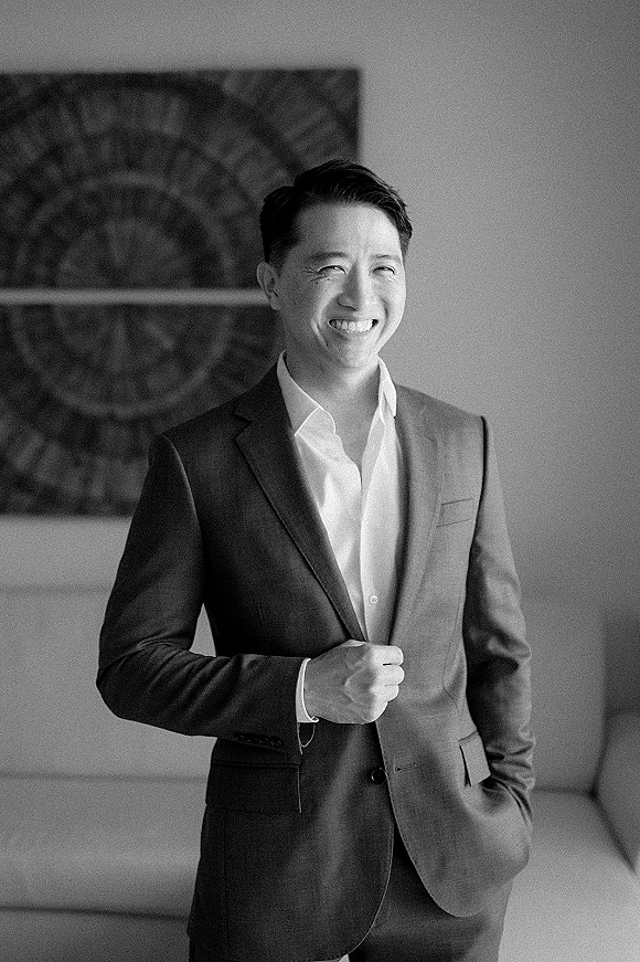 Groom portrait of a man in a dark suit with pocket square, standing against an interior wall with framed artwork in the background