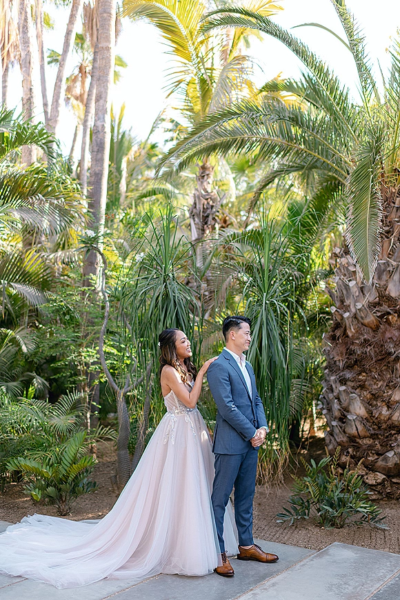First look moment as the bride taps the groom’s shoulder on a stone walkway in a tropical garden, her long dress train trailing behind