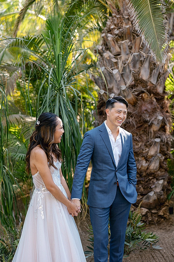 Couple portrait of a laughing bride and groom holding hands, her lace wedding dress and his blue suit amid palm trees and lush garden greenery